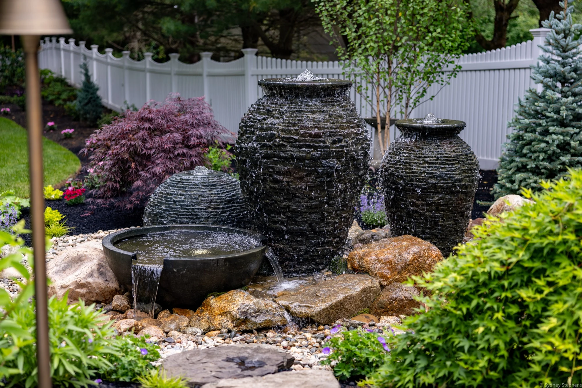 Water spilling down two stacked slate urns and a stacked slate sphere into a black spillway bowl that pours over fieldstone, framed by a Japanese maple and blue spruce