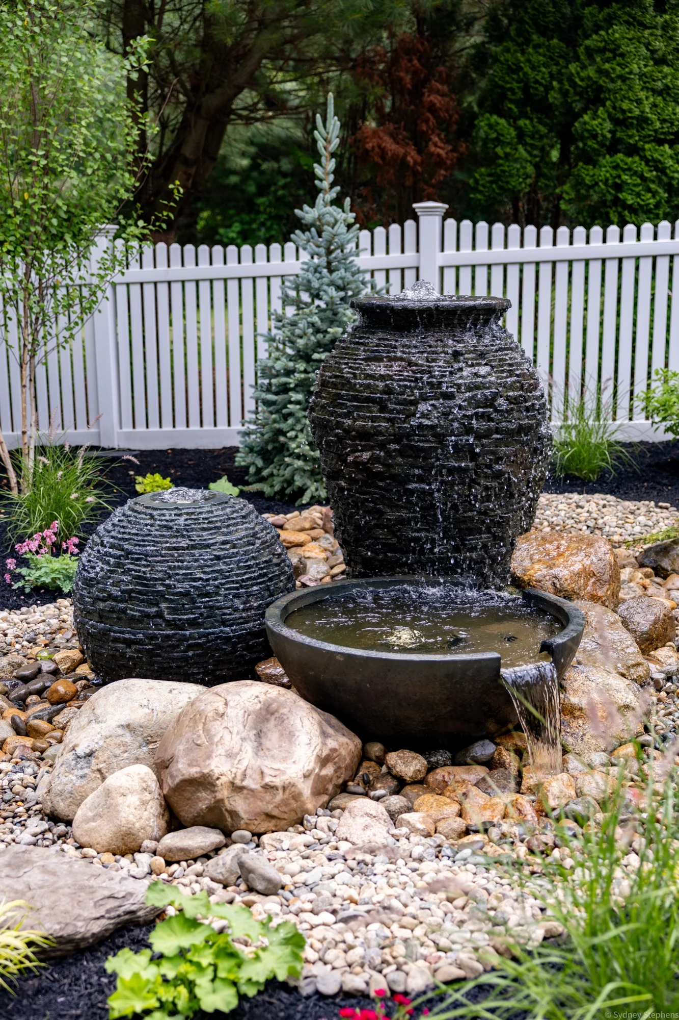 Tall stacked slate urn and stacked slate sphere behind a black spillway bowl pouring onto river rock, with a blue spruce and white picket fence behind